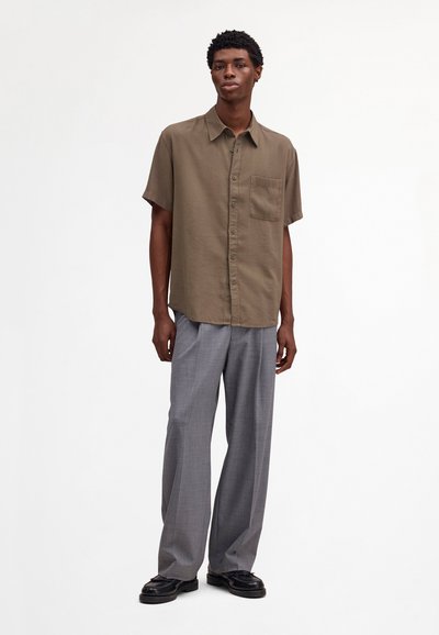 Jeune homme debout, portant une chemise marron à manches courtes avec boutons, un pantalon gris à jambes larges et des mocassins noirs, sur fond blanc.