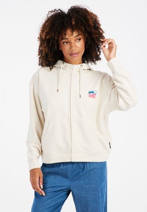 Cream-colored zip-up hoodie with a front pocket, hood, and drawstrings. Features a small colorful logo patch on the chest. Soft fabric texture.
