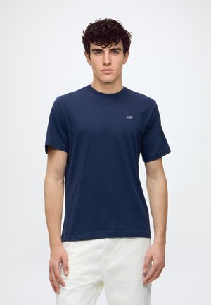 Jeune homme aux cheveux bouclés portant un t-shirt bleu marine et un pantalon blanc, debout devant un fond clair uni.