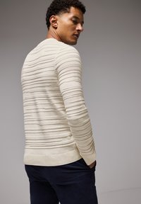 Krämfärgad tröja med ribbat mönster, långa ärmar och en figurnära design. Bärs över mörka byxor, visas från en bakvy.