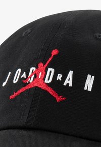 Melnā kokvilnas cepure ar noapaļotu vainagu. Uz priekšu ir balts "JORDAN" teksts un sarkans izšuvēts Jumpman logo.