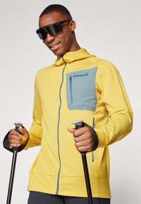 Yellow zip-up hoodie with light blue pocket, textured fabric, and adjustable hood. Model holds trekking poles. Glasses frame rounded shape.