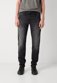 Personne portant un jean noir slim délavé, des baskets noires et un t-shirt gris, debout devant un fond blanc uni.