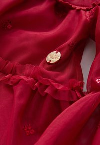Tessuto rosso trasparente con rifiniture a balze, piccoli dettagli floreali ricamati e un bottone rotondo dorato con incisione floreale.