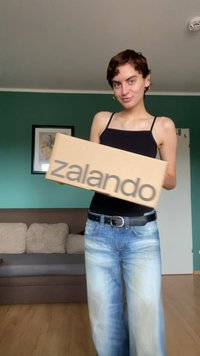 Zalando kartona kaste, ko tur rokās persona, ar pelēku logotipu uz brūnas virsmas. Redzami arī ikdienišķi zili džinsi un melna blūze.