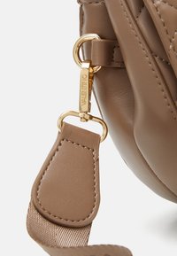 Barna bőr kézitáska sarkán arany hardware, kulcskarikával és texturált pánttal. A pánton látható a varrás részlete.