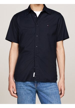 Uomo che indossa una camicia blu navy a maniche corte con bottoni sopra una canottiera bianca e jeans blu, in piedi con le braccia rilassate.