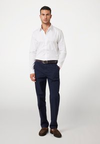 Camisa blanca de botones con mangas largas, pantalones navy y mocasines marrones texturizados, llevada con un cinturón marrón, de pie contra un fondo neutro.
