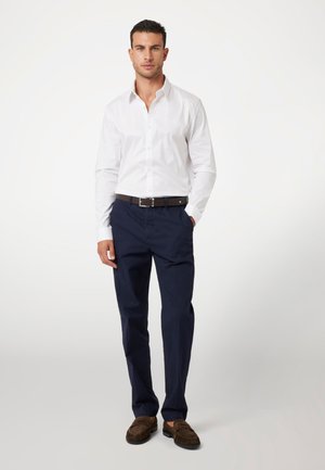 Camisa blanca de botones con mangas largas, pantalones navy y mocasines marrones texturizados, llevada con un cinturón marrón, de pie contra un fondo neutro.