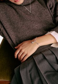 Grauer gestrickter Pullover, getragen über einem weißen Hemd mit Kragen, mit einem silbernen Gliederarmband, das einen kleinen Anhänger zeigt, auf einer Hand, die auf einem dunklen Stoff ruht.