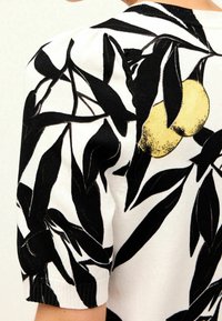 Biała tkanina z czarnym liściastym wzorem i dwoma wydrukowanymi żółtymi cytrynami blisko ramienia na krótkiej, puffowanej rękawie.