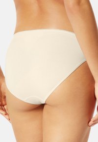 Frau trägt beige, hohe Baumwollslips, von hinten gezeigt mit entspannten Händen an den Seiten, auf hellgrauem Hintergrund.