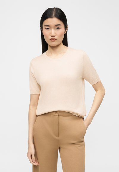 Γυναίκα με ίσια μαύρα μαλλιά φοράει ανοιχτό μπεζ μπλουζάκι με κοντά μανίκια, μπαίνει μέσα σε ψηλόμεσο παντελόνι καμηλό χρώματος, με το χέρι στην τσέπη.