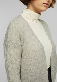 Ljust grå stickad cardigan med V-halsdesign och texturerade, mönstrade ärmar. Bärs över en åtsittande krämfärgad polotröja. Mjuk, fluffig textur.