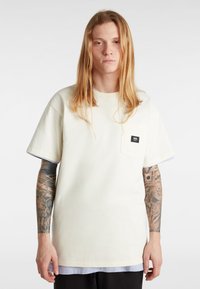 Camiseta de color beige claro con cuello redondo, mangas cortas y un bolsillo en el pecho. Presenta una etiqueta de marca negra en el bolsillo. Tejido de algodón suave.