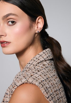 Teksturowany beżowy bezrękawnik z wysokim kołnierzem. Modelka nosi małe niebieskie kolczyki wkrętki. Gładkie włosy ułożone w niskiego kucyka.