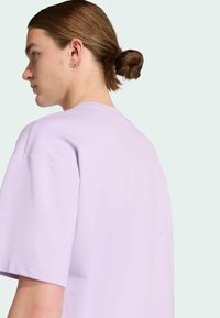 Persona joven con el pelo castaño recogido en un moño, vistiendo una camiseta de manga corta lisa color lavanda, mirando hacia la izquierda con un fondo neutro.