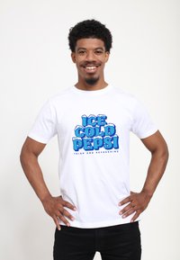 Bílý tričko s krátkým rukávem s výrazným modrým textem "ICE COLD PEPSI" s ledovými akcenty, které má na sobě osoba s kudrnatými vlasy, se založenýma rukama v bocích.