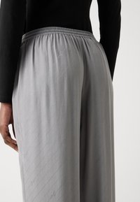 Grå elastiske bukser med strikk i livet, med diagonale striper og subtile logomønstre. Stoffet virker glatt med en lett tekstur.