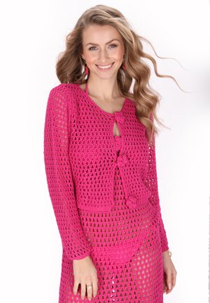 Copertura in crochet rosa con dettaglio a nodo sul davanti, maniche lunghe, motivo a rete aperta e una texture leggermente trasparente.