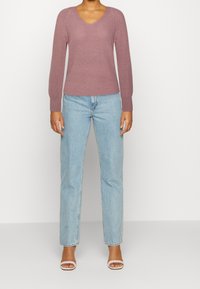 Maglione a V in maglia color malva con maniche lunghe e polsini a costine, abbinato a jeans dritti blu chiaro e sandali bianchi con tacco.