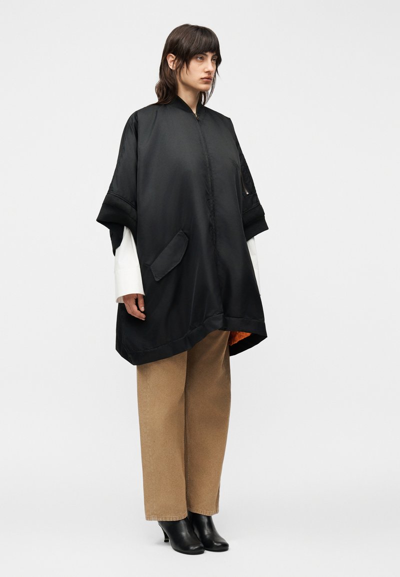 Femme portant une veste noire ample à manches courtes sur une chemise blanche à manches longues, pantalon large couleur beige et bottines noires, debout devant un fond blanc.