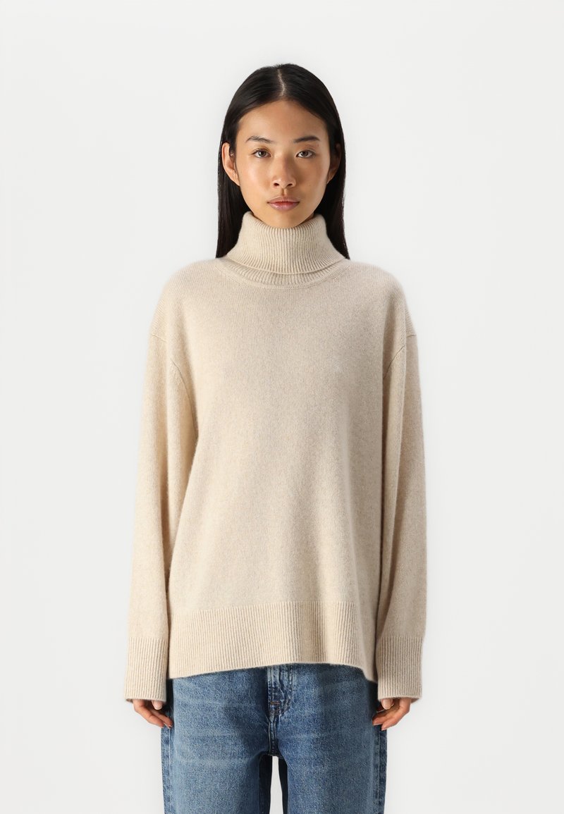 Pull à col roulé couleur crème avec une texture côtelée, coupe ample et épaules tombantes, associé à un jean en denim bleu.