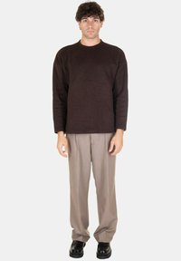 Maglione marrone scuro abbinato a pantaloni grigi chiari con pieghe. L'outfit presenta una vestibilità rilassata, contrasto di texture e scarpe nere.