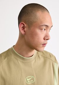 Krátký rukáv košile v odstínu tan s logem Nike Air v zelené barvě, kulatým výstřihem a kontrastním světle zeleným lemováním výstřihu. Detailní boční profil.