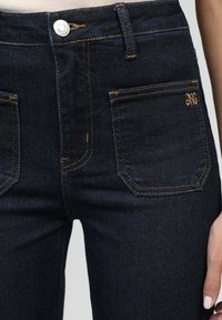 Zoom sur un jean en denim bleu foncé montrant les poches avant, des coutures orange, une fermeture à bouton, et une partie d'une main avec du vernis à ongles rose pâle.