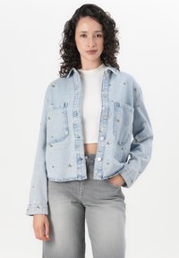 Lyseblå denimjakke med broderte blomstermotiver, knappeåpning, to frontlommer og en cropped design. Brukt over en hvit topp.