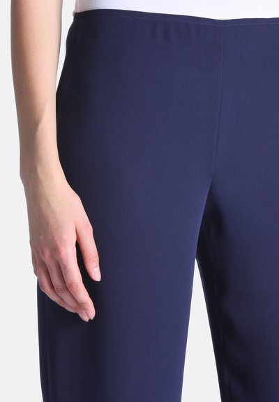 Gros plan d'une personne portant un pantalon bleu marine ajusté avec une taille lisse et une main reposant à côté de la cuisse.