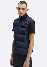 Giovane uomo con indosso un gilet trapuntato blu navy senza maniche sopra una t-shirt nera, in piedi davanti a uno sfondo chiaro uniforme.