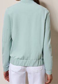 Kobieta ubrana w jasnoniebieską bluzkę z długim rękawem i elastycznym dołem oraz białe spodnie, przedstawiona od tyłu na neutralnym tle.