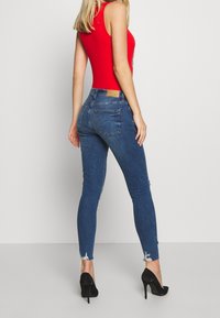 Jeans de denim azul ajustados con dobladillo deshilachado, combinados con una blusa roja sin mangas ceñida y zapatos de tacón alto negros.