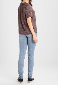 Frau trägt ein gemustertes kurzärmliges Oberteil, hellblaue skinny Jeans und schwarze Turnschuhe, steht leicht abgewandt vor einem einfarbigen Hintergrund.