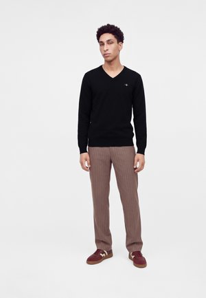 Jeune homme debout, portant un pull noir col en V, un pantalon marron à fines rayures et des baskets bordeaux avec des touches beige, sur un fond clair uni.