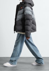 Svart och grå puffjacka med huva, med en orange logotyp, kombinerad med ljusblå vida jeans och vita sneakers.