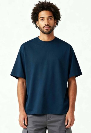 Jeune homme aux cheveux bouclés portant un t-shirt bleu marine oversized et un pantalon cargo gris, debout devant un fond blanc uni.