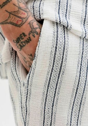 Main tatouée avec le texte "SUPER SAIYAN" partiellement visible dans une poche de vêtement blanc à rayures verticales noires.