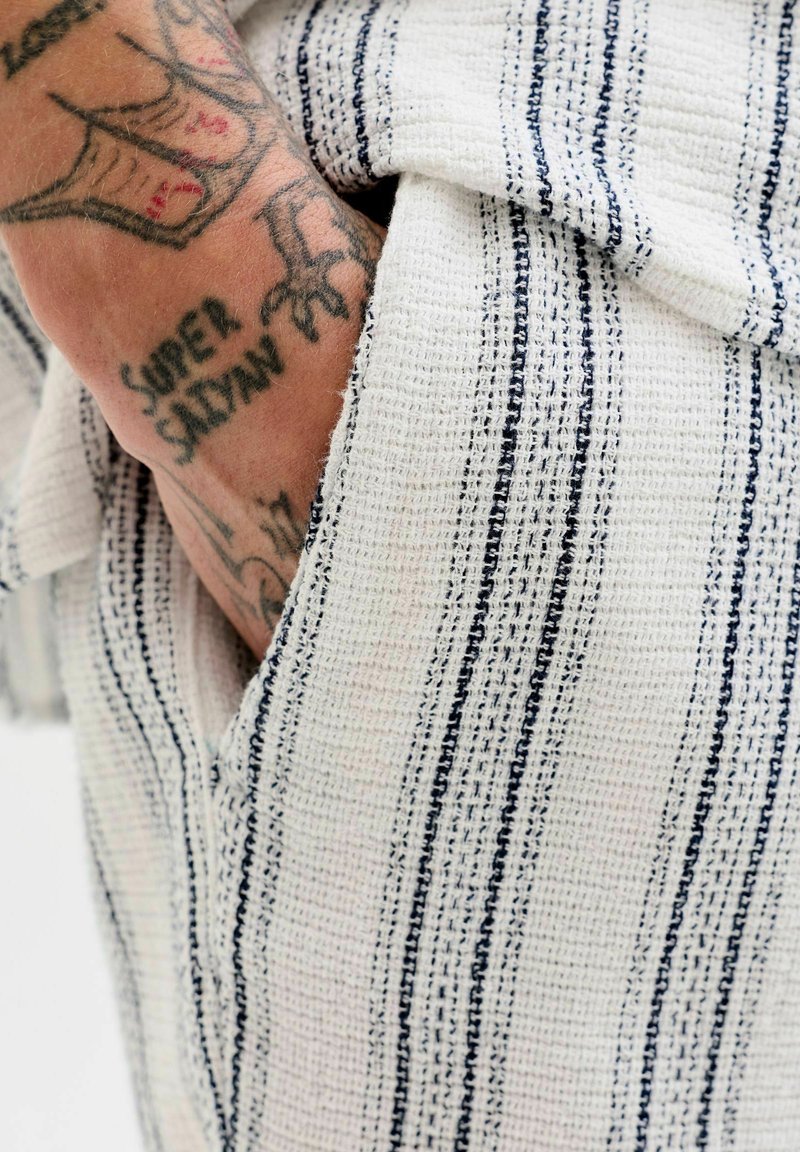 Main tatouée avec le texte "SUPER SAIYAN" partiellement visible dans une poche de vêtement blanc à rayures verticales noires.