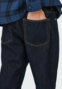Jean en denim bleu foncé avec des coutures orange, comportant des poches arrière et une coupe droite, partiellement affiché aux côtés d'une chemise à carreaux.