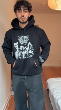 Corta-vento preto com uma gráfica frontal de design branco em contraste; apresenta um bolso canguru e punhos canelados; combinado com jeans escuros.
