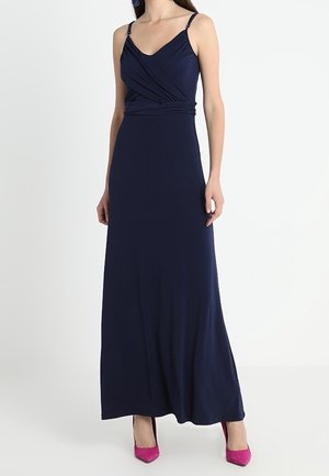 Kobieta ubrana w granatową, bezrękawnikową suknię maxi z kopertowym stanikiem i różowymi butami na wysokim obcasie, stojąca na tle jednolitego białego tła.