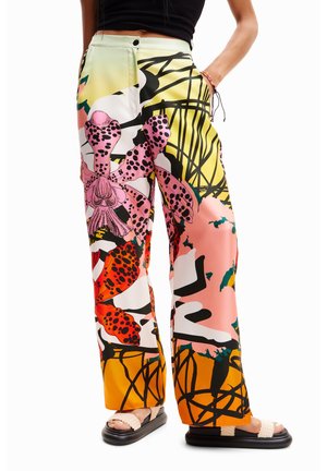 Femme portant un pantalon à jambes larges avec de grands motifs floraux colorés en rose, orange, jaune et noir, associé à des sandales beiges et un crop top noir.