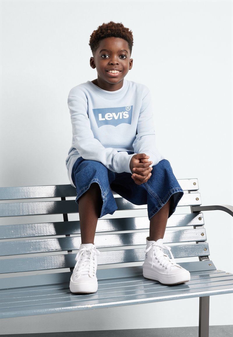 Sudadera azul clara con logo blanco de Levi's, combinada con pantalones cortos de denim azul hasta la rodilla y zapatillas altas blancas, sentado en un banco metálico.