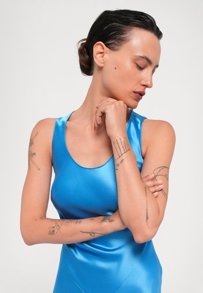 Satijnen blauwe tanktop met een gladde, glanzende textuur en ronde halslijn, gedragen door een persoon met zichtbare tatoeages, met gekruiste armen.