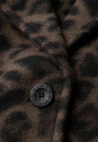 Leopardenmusterstoff in Brauntönen und Schwarz, mit einem runden schwarzen Knopf mit vier Löchern, aus strukturiertem Wollmaterial.