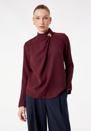 Mujer con blusa de manga larga color burdeos con detalle drapeado y broche dorado, combinada con pantalones anchos de rayas marineras sobre fondo blanco.