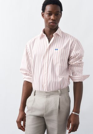 Νέος άνδρας που φοράει ανοιχτό ροζ πουκάμισο με λεπτές ρίγες και μανίκια γυρισμένα, καθώς και μπεζ σαλοπέτες, στέκεται μπροστά σε λευκό φόντο.